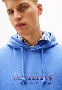 Felpa blu con cordini presenta un logo "tommy" multicolore. Sotto, una maglietta bianca con strisce blu e grigie è parzialmente visibile.