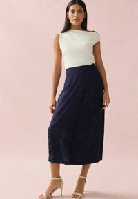Top bianco off-the-shoulder abbinato a una lunga gonna plissettata blu navy con un motivo floreale. La modella indossa tacchi a spillo beige con cinturino alla caviglia.