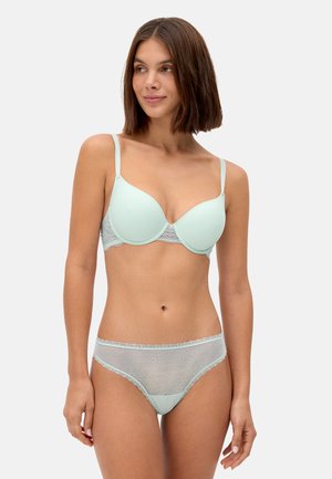 Mujer con cabello corto y castaño, vestida con sujetador acolchado azul claro y ropa interior de encaje transparente a juego, posando frente a un fondo liso.