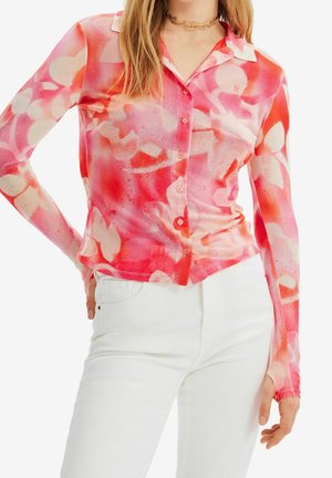 Frau trägt eine enganliegende, langärmelige, knöpfbare Bluse mit pinken und roten Blumenmustern, kombiniert mit weißen Hosen.