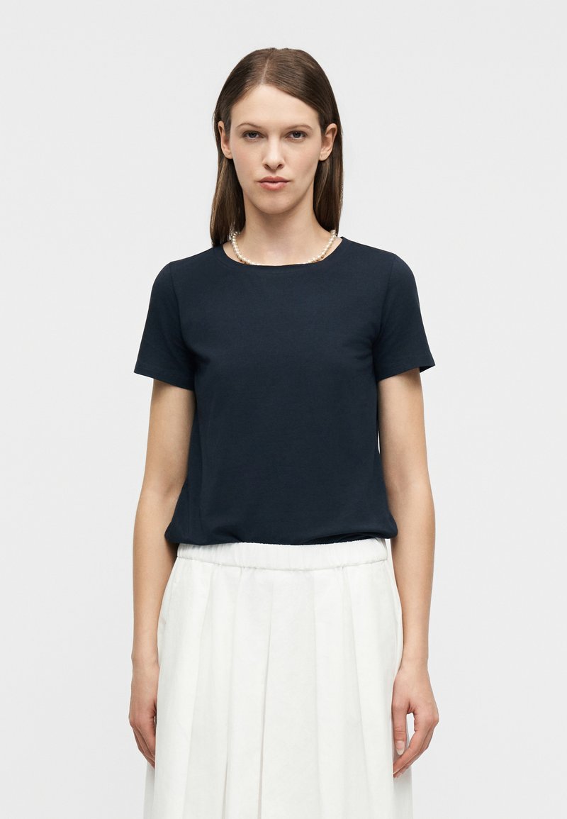 Γυναίκα με ίσια καστανά μαλλιά, που φοράει μπλε ναυτικό κοντομάνικο πουκάμισο, λευκή πλισέ φούστα και περιδέραιο από πέρλες, στέκεται μπροστά σε λευκό φόντο.