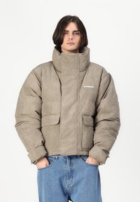 Duvet jakna od teksturiranog bež materijala s visokim ovratnikom, prednjim džepovima i detaljem loga. Uparena s širokim plavim trapericama.