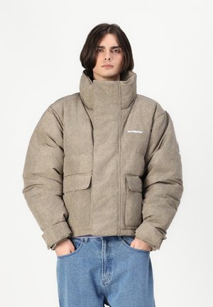 Puffer bunda v texturovaném béžovém materiálu s vysokým límcem, předními kapsami a detailním logem. V kombinaci se volnými modrými džíny.