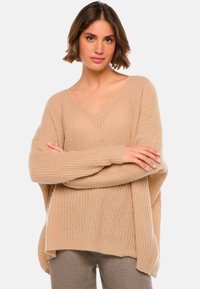 Pull en tricot beige avec un décolleté en V profond, coupe loose et manches longues. Le tissu texturé présente un motif côtelé.