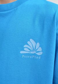 Camiseta de algodón azul claro con cuello redondo y un sutil gráfico de logo blanco en la parte frontal. Textura suave con mangas cortas.