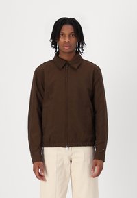 Veste marron à fermeture éclair avec col, poches avant et taille élastique, en tissu lisse. Modèle portant un pantalon de couleur claire.