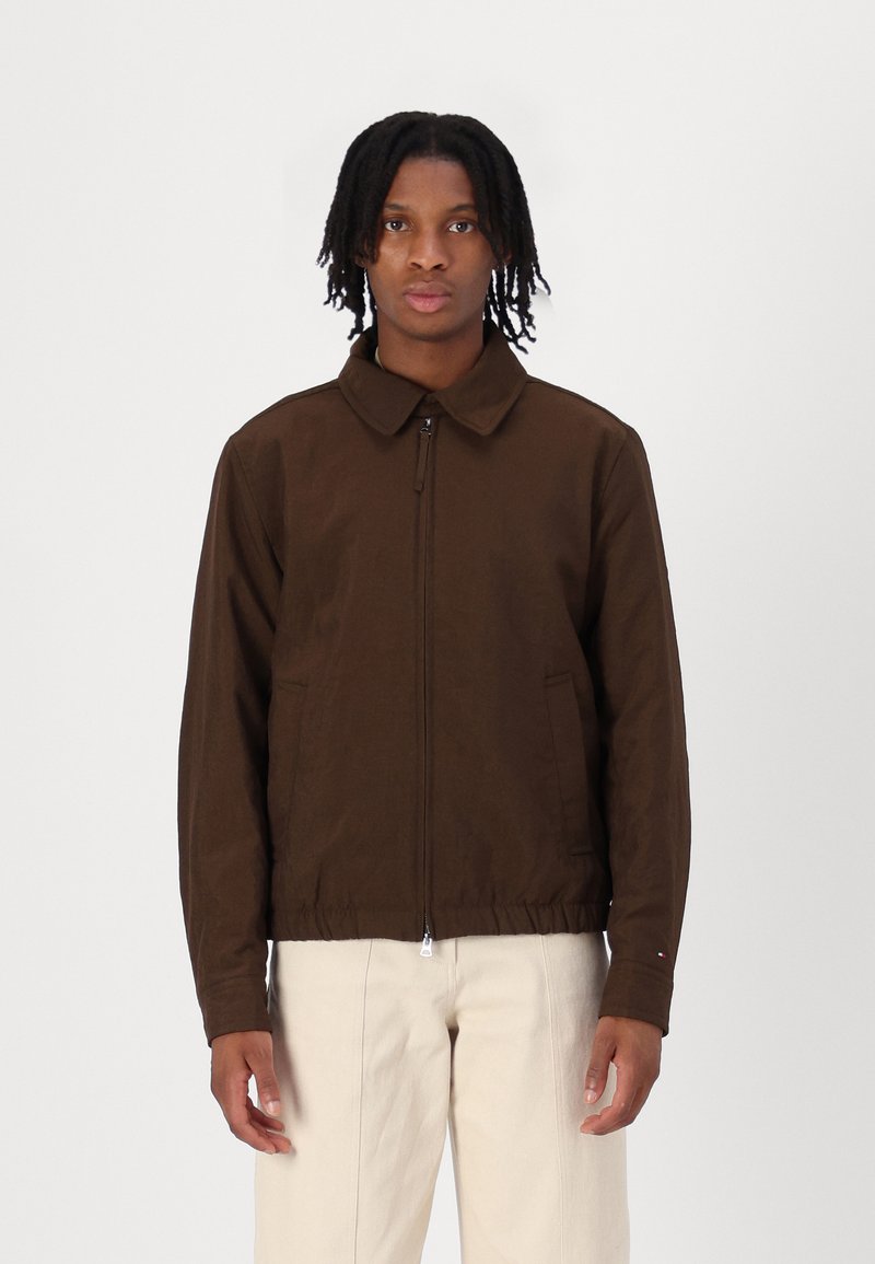 Veste marron à fermeture éclair avec col, poches avant et taille élastique, en tissu lisse. Modèle portant un pantalon de couleur claire.