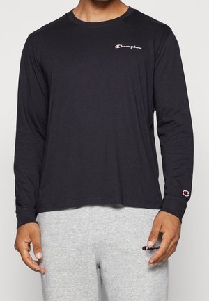 Uomo che indossa una maglia nera Champion a maniche lunghe e pantaloni della tuta grigio chiaro con loghi Champion visibili sul petto, sulla manica e sui pantaloni.