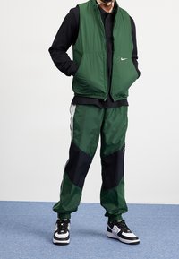 Man som bär en grön zip-up väst och matchande gröna och svarta nylon joggingbyxor, svart skjorta och svart-vita Nike sneakers, står på ett blått golv.