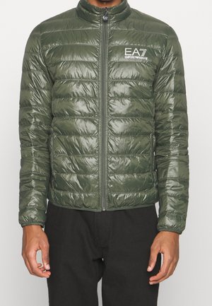Homme portant une doudoune vert olive brillante avec une fermeture éclair devant et le logo blanc EA7 Emporio Armani sur la poitrine.