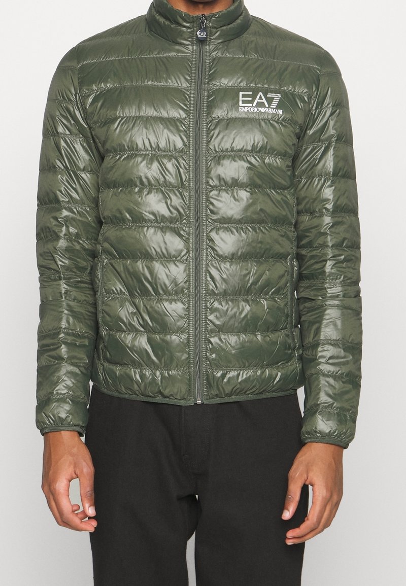 Homme portant une doudoune vert olive brillante avec une fermeture éclair devant et le logo blanc EA7 Emporio Armani sur la poitrine.