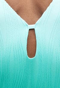 Κοντινό πλάνο ενός ατόμου που φοράει υφασμάτινο τουρκουάζ τοπ με κεντρικό οπτικό άνοιγμα μπροστά, στερεωμένο με οριζόντια υφασμάτινη πόρπη.