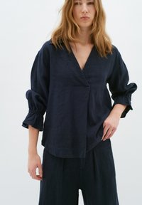 Top in lino blu navy con scollo a V, maniche lunghe a palloncino con polsini elasticizzati e vestibilità morbida e drappeggiata. È visibile anche la combinazione con pantaloni coordinati.