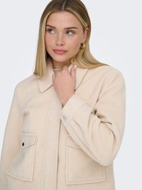 Ung kvinde med blondt hår iført guld øreringe i hoop-stil og en beige uldfrakke, der holder i frakkens krave med den ene hånd.