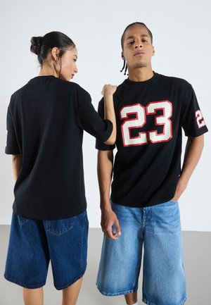 T-shirts noirs avec un design du numéro rouge "23", associés à des shorts en jean—en délavé foncé et clair. Le modèle pose dos à dos sur un fond neutre.