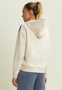 Felpa con cappuccio beige chiaro, dal taglio morbido, con polsini e bordo a costine. Presenta un tessuto testurizzato e un logo discreto sulla manica.