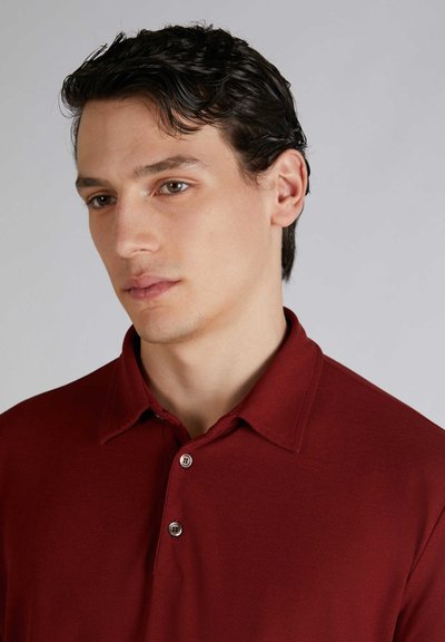 Polo di colore bordeaux con colletto, dotato di pattina con tre bottoni, texture liscia e maniche corte. Ingrandimento della parte superiore del viso di un modello.