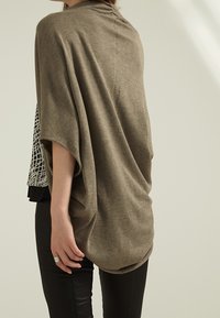 Brązowy dziergany ponczo z rękawami nietoperzowymi, o fakturowanym wykończeniu i nieregularnym dole. Połączony z dopasowanymi czarnymi spodniami.