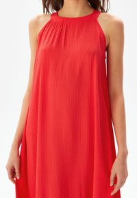 Robe rouge sans manches avec une texture côtelée et un décolleté froncé. Silhouette fluide, longueur genoux, tissu léger. Pas de motifs ou d'accents supplémentaires.