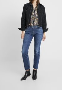 Zwarte gewatteerde jas met drukknoopsluitingen, gestreepte blouse, blauwe jeans en zwarte puntige enkellaarsjes. Glad textiel en levendige kleuren.