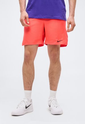 Mann trägt leuchtend rote Nike-Shorts, weiße Nike-Sneaker und ein violettes Shirt, steht vor einem schlichten weißen Hintergrund.