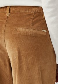 Bruine corduroy broek met verticale ribbels en een enkele achterzak. De stof heeft een zachte textuur en een subtiele glans.