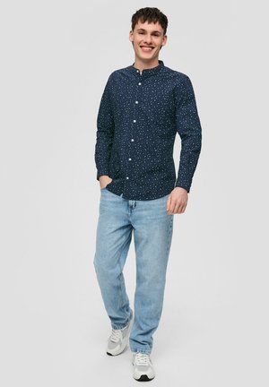 Marineblauwe overhemd met kleine witte stippen, lange mouwen en een mandarinakraag; gecombineerd met lichtblauwe jeans en witte sneakers.