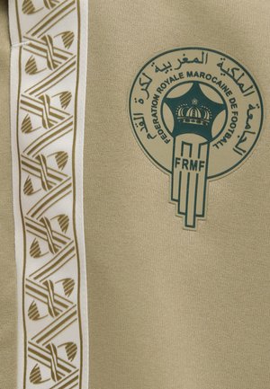 Indumento beige con il logo della Fédération Royale Marocaine de Football e una striscia verticale bianca con motivi geometrici dorati.