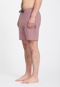 Homme debout, pieds nus, portant un short de bain mauve avec un cordon noir et un logo noir et blanc sur la cuisse droite, sur fond blanc.