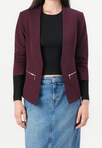 Blazer bordeaux à coupe ajustée, deux poches avant zippées et manches trois-quarts, associé à un haut noir et une jupe en jean.