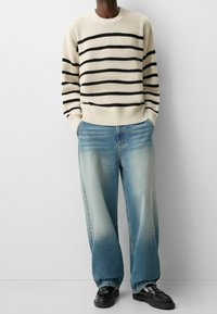 Suéter de punto crema con rayas negras, combinado con jeans de pierna ancha en azul claro, que difuminan de oscuro a claro. Mocasines negros completan el conjunto.