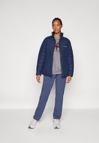 Giacca isolante blu navy con colletto alto, indossata sopra una maglietta grigia con grafica, abbinata a pantaloni blu e sneakers grigie.
