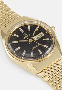 Montre en quartz dorée avec un cadran noir, des index horaires blancs, un affichage de la date et un bracelet en maille métallique à motif. Présente une finition polie.