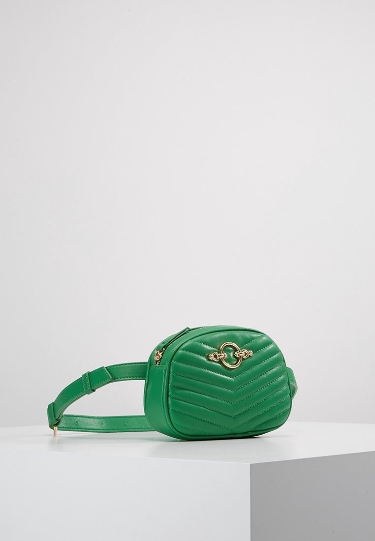 Petite sacoche ceinture en cuir vert avec surpiqûres en chevron et fermoir circulaire doré, présentée sur une surface blanche contre un fond uni.