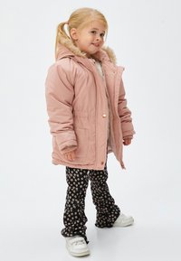 Niña joven con cabello rubio en una coleta, vestida con un abrigo rosa con capucha y borde de piel, pantalones negros con flores y zapatillas blancas, de pie y sonriendo.
