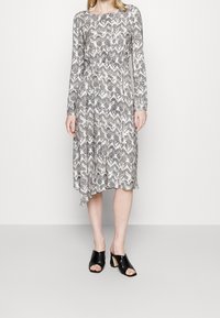 Šaty s dlouhým rukávem s černo-bílým abstraktním vzorem listů, se staženým pasem a asymetrickým lemem. Nosí se s černými sandály na vysokém podpatku s otevřenou špičkou.