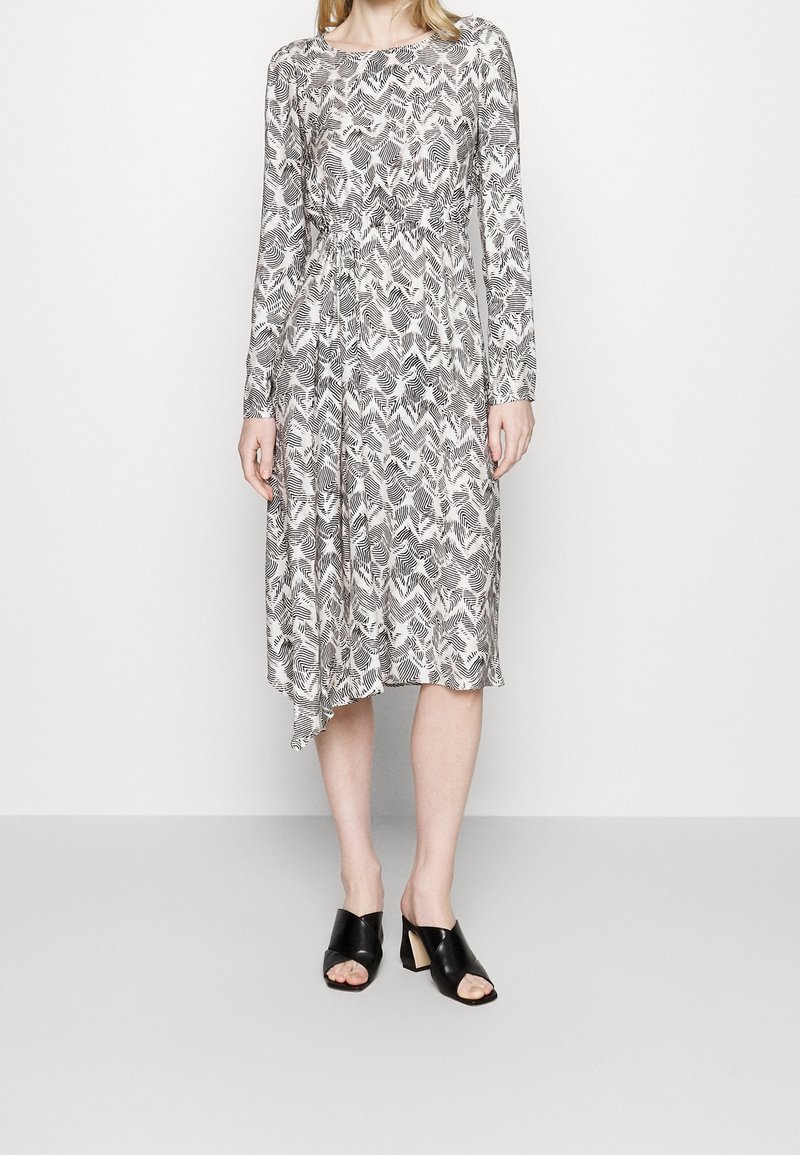 Šaty s dlouhým rukávem s černo-bílým abstraktním vzorem listů, se staženým pasem a asymetrickým lemem. Nosí se s černými sandály na vysokém podpatku s otevřenou špičkou.