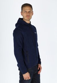 Felpa con zip di colore blu navy, realizzata in tessuto testurizzato, con cappuccio, due tasche laterali con zip e un piccolo logo sul petto.