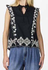 Haut noir sans manches avec une broderie florale blanche le long des bords et du décolleté, avec un col noué et un ourlet festonné.