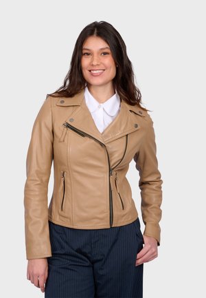 Smiling woman wearing a beige leather zip-up jacket over a white collared shirt and dark pinstriped pants, standing against a plain background.