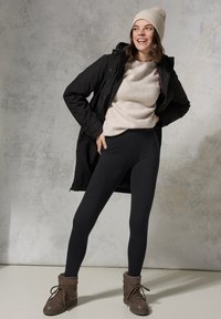 Zwarte leggings gecombineerd met een beige geribde trui onder een zwarte jas, aangevuld met bruine veterlaarzen en een bijpassende beanie.