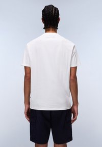 Camiseta de algodón blanca de manga corta con cuello redondo, vista desde la parte posterior. Presenta un diseño limpio y un corte relajado, combinada con pantalones cortos azul marino.