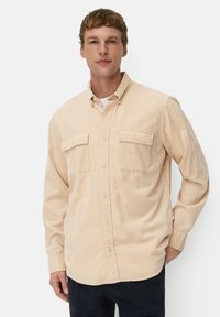 Beige langemouwen overhemd met knopen en twee borstzakken, zachte stof; gedragen met een wit ronde hals T-shirt eronder.