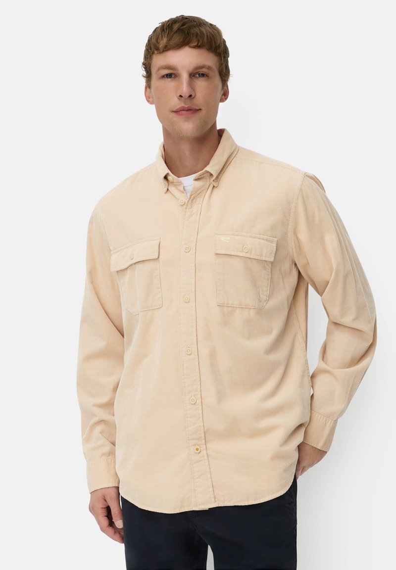 Beige langemouwen overhemd met knopen en twee borstzakken, zachte stof; gedragen met een wit ronde hals T-shirt eronder.