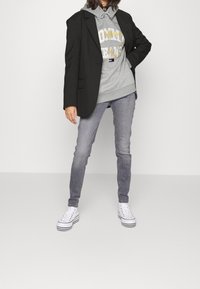 Blazer nero sopra un hoodie grigio con testo, jeans grigi e sneakers alte bianche con strisce nere. Tessuti morbidi e vestibilità rilassata.