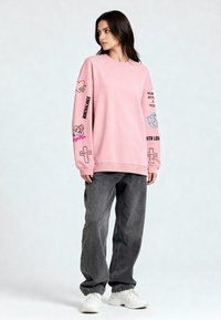 Rózsaszín, oversized kapucnis pulcsi, amelyen különböző nyomtatott részletek találhatóak az ujjakon, szürke, bő szabású farmernadrággal és fehér sneaker cipőkkel párosítva. Simán tapintású, laza szabású.