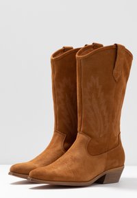 Brązowe zamszowe kowbojki z dekoracyjnym haftem w liście, spiczastymi czubkami i niskim, obcasem na koturnie. Wyposażone w pętle do łatwego zakupu.