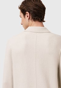 Pull en maille couleur crème avec col, présentant une surface texturée et un détail de couture au dos. Boucle d'oreille argentée visible sur le modèle.
