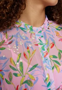 Blouse rose transparente avec un motif floral agrémenté d'accents verts et rouges, col rond et devant à boutons avec de subtils détails métalliques.
