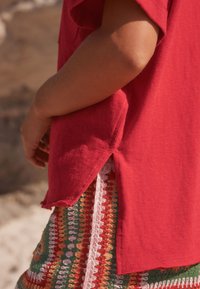 T-shirt rouge avec une fente latérale, doté d'un ourlet effiloché et d'une texture douce, superposé à une jupe à motifs multicolores avec des accents crochetés.